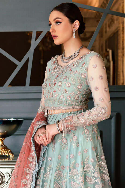 ''Woman in a light blue embroidered traditional outfit with pink dupatta against a decorative wall.''