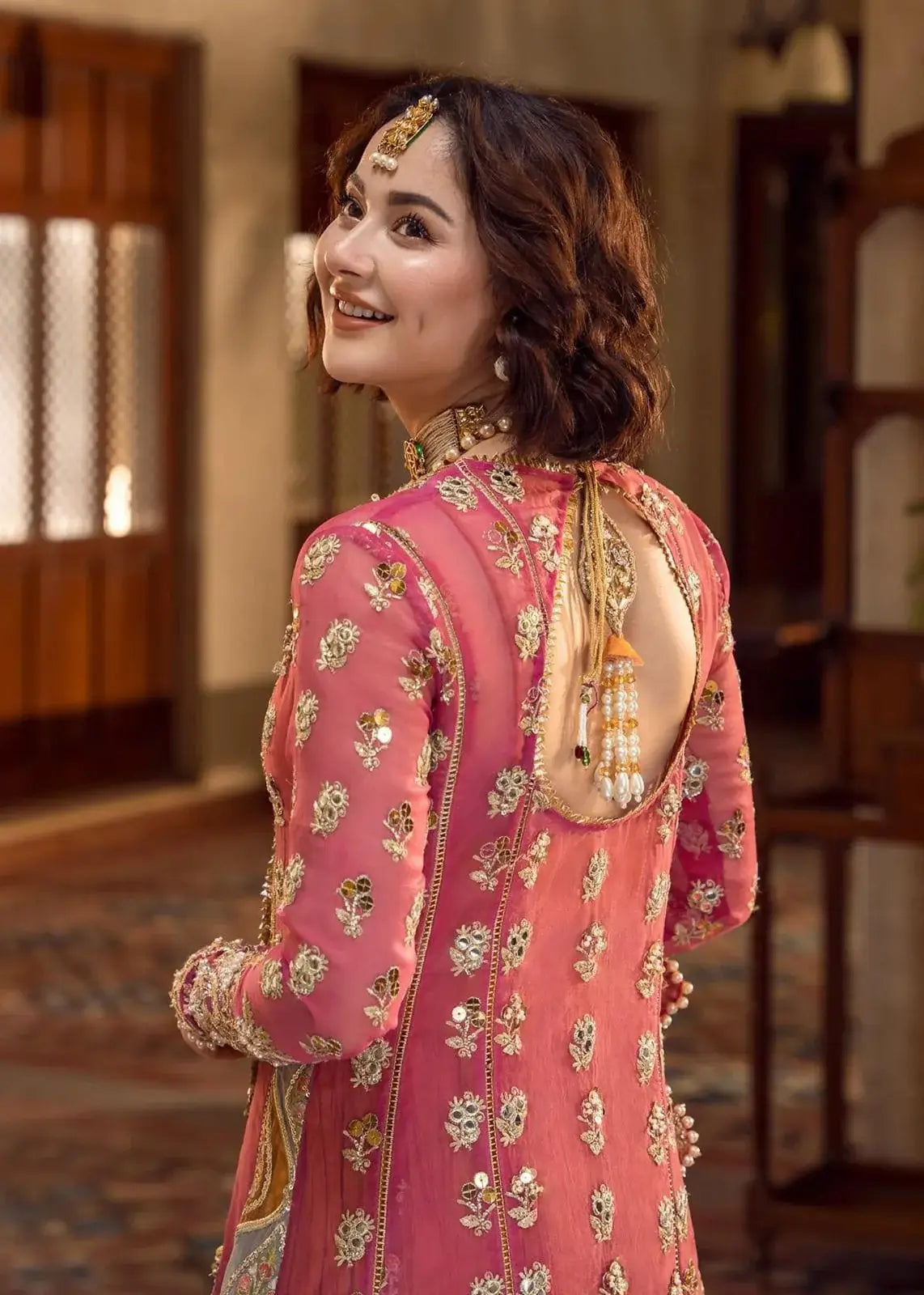 ''Woman in a pink traditional outfit with floral patterns and gold accents, standing indoors.''